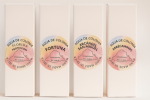 Cuatro cajas blancas de Agua de Colonia artesanal de la marca Creaciones Es Vedrà, alineadas sobre un fondo claro. Cada caja tiene una etiqueta circular con tonos pastel y los nombres de las fragancias: “Florida Protección”, “Fortuna”, “Arcángel San Miguel” y “Abrecaminos”. En las etiquetas se lee “Hand Made in Ibiza”.