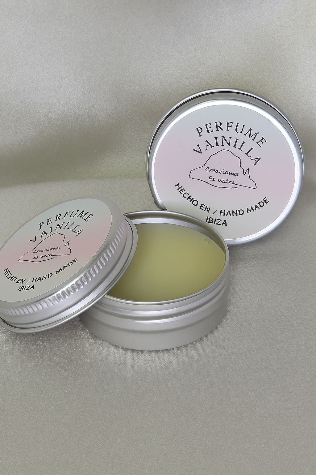Perfume sólido con aroma a vainilla de Creaciones Es Vedra, presentado en envase metálico plateado con tapa rosa y texto “Hecho a mano en Ibiza”. Muestra el producto abierto sobre fondo suave, destacando su textura cremosa y estilo artesanal.