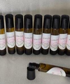 Fotografía de varios frascos de Roll-On Holístico Amor alineados sobre fondo claro. Los envases de vidrio ámbar con etiqueta rosa y negra muestran la marca “Creaciones Es Vedrà” y el texto “Hecho a mano en Ibiza”. Un frasco aparece abierto en primer plano mostrando el aplicador tipo roller.