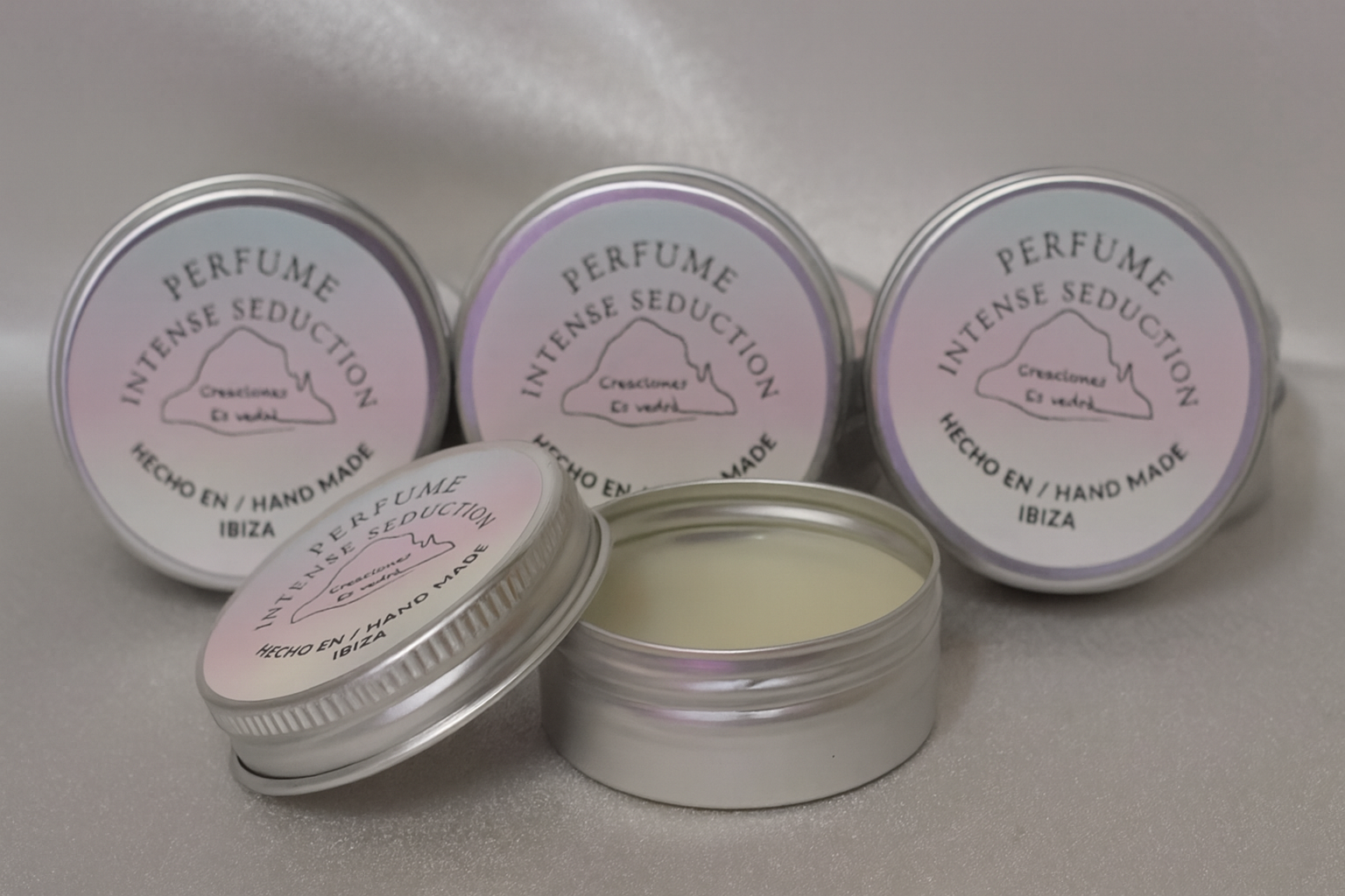 Cuatro latas plateadas de perfume sólido artesanal "Intense Seduction" de Creaciones Es Vedrà, hechas a mano en Ibiza. Una de las latas está abierta mostrando la textura cremosa del perfume, colocadas sobre un fondo suave y satinado.
