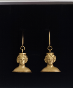 Par de pendientes dorados inspirados en la Diosa Tanit, con diseño artesanal en forma de busto clásico, presentados sobre fondo negro en caja de joyería.