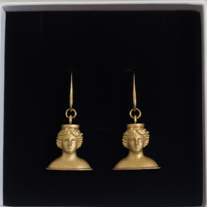 Par de pendientes dorados inspirados en la Diosa Tanit, con diseño artesanal en forma de busto clásico, presentados sobre fondo negro en caja de joyería.